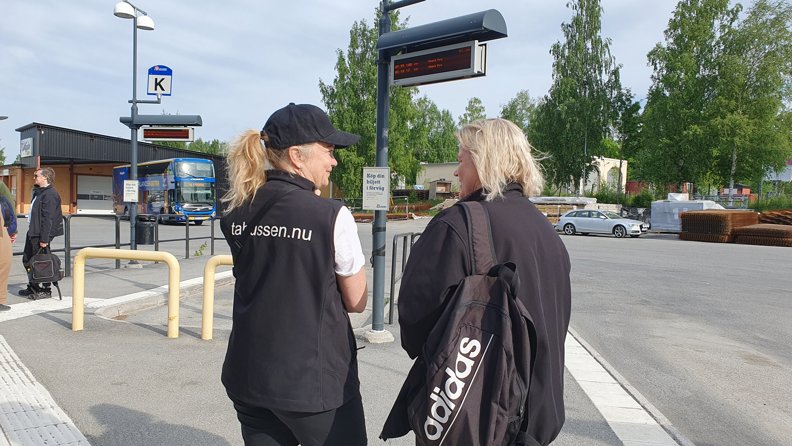 Två personer samtalar på Skellefteå busstation, digitala hållplatsskyltar syns i bakgrunden